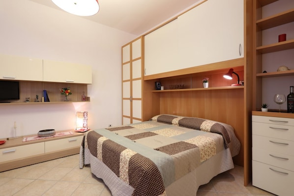 A bedroom with a bed, a desk, a TV, and a shelf with decorative items.