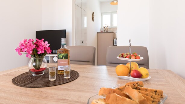 Un tavolo da pranzo con un vaso di fiori rosa, una bottiglia di vodka Smirnoff, un bicchiere d'acqua, un piatto di frutta e un piatto di grissini.