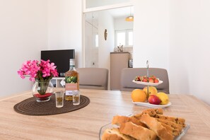 A dining table with a vase of pink flowers, a bottle of Smirnoff vodka, a glass of water, a plate of fruit, and a plate of breadsticks. - Apartment Oleander (Punat)