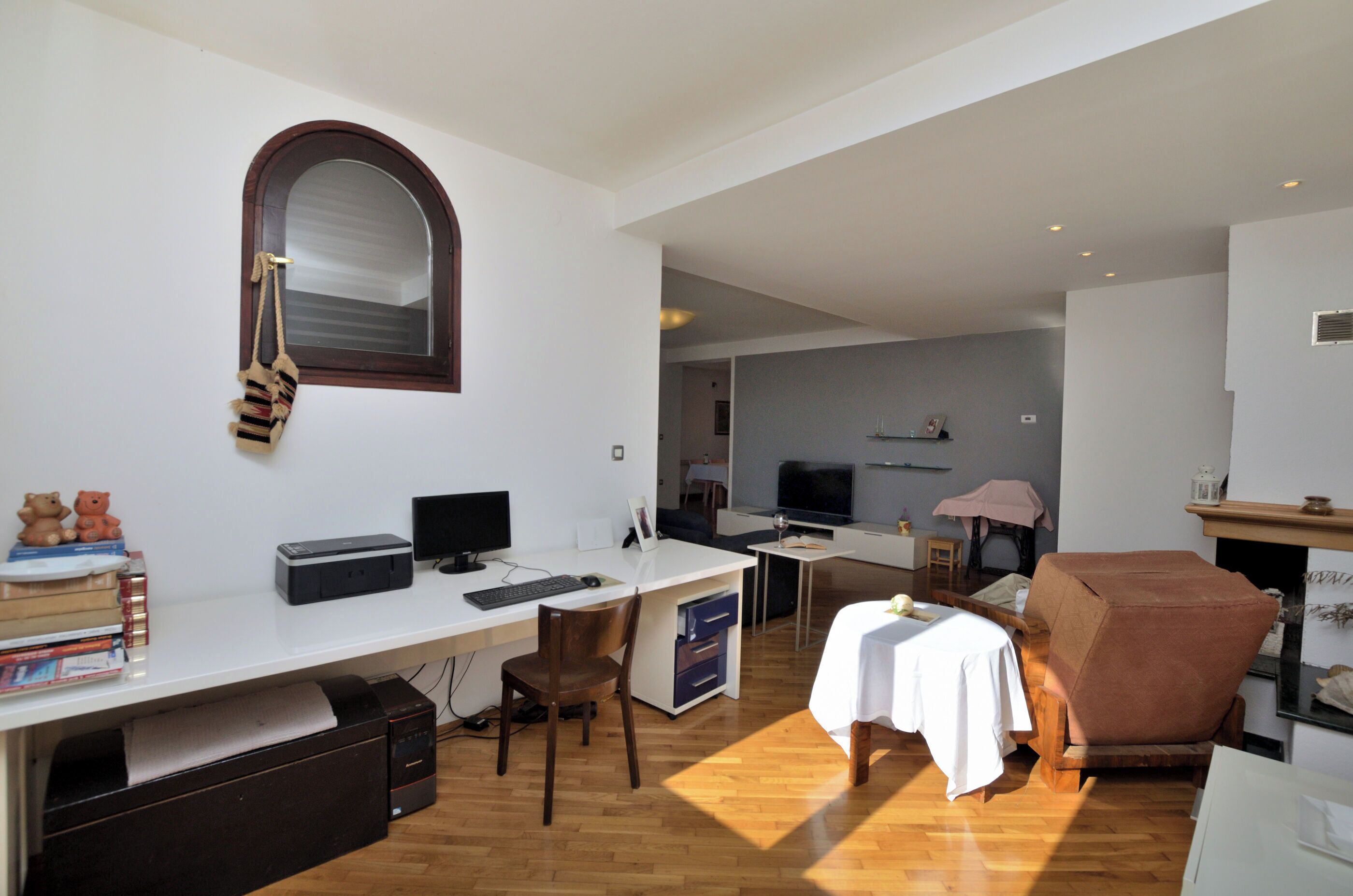 Een moderne woonkamer met een wit bureau, een computer en een televisie. Er staat een bruine fauteuil en een klein tafeltje met een witte tafelkleed.