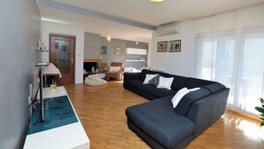 Ein modernes Wohnzimmer mit einem Ecksofa, einem Couchtisch und einem Flachbildfernseher. Holzfußboden und eine Tür zu einem weiteren Raum.