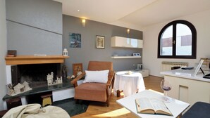 Ein gemütliches Wohnzimmer mit Kamin, einem bequemen Sessel, einem kleinen Tisch mit Buch und einem Glas Wein sowie einem großen Bogenfenster.
