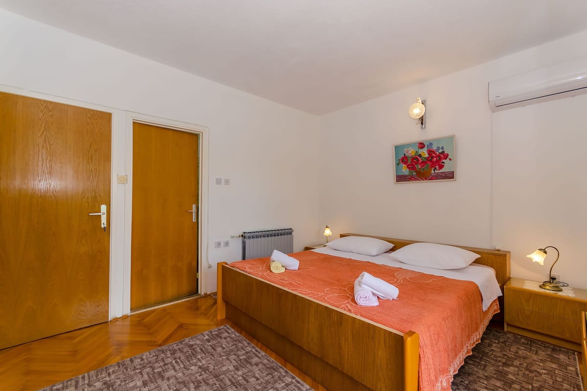 A bedroom with a wooden bed, white walls, a radiator, a painting, and a nightstand with a lamp.