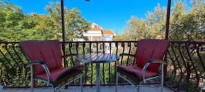 A balcony with two red chairs and a small round table, surrounded by a metal railing, overlooking a residential area with trees and a house. - Rooms Ante (Jelsa)
