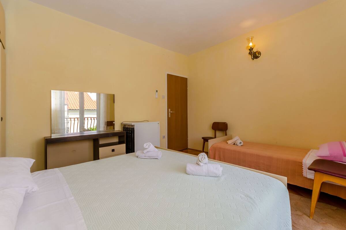 A hotel room with a large bed, a desk with a mirror, a small table, a chair, and a single bed with a pink pillow.