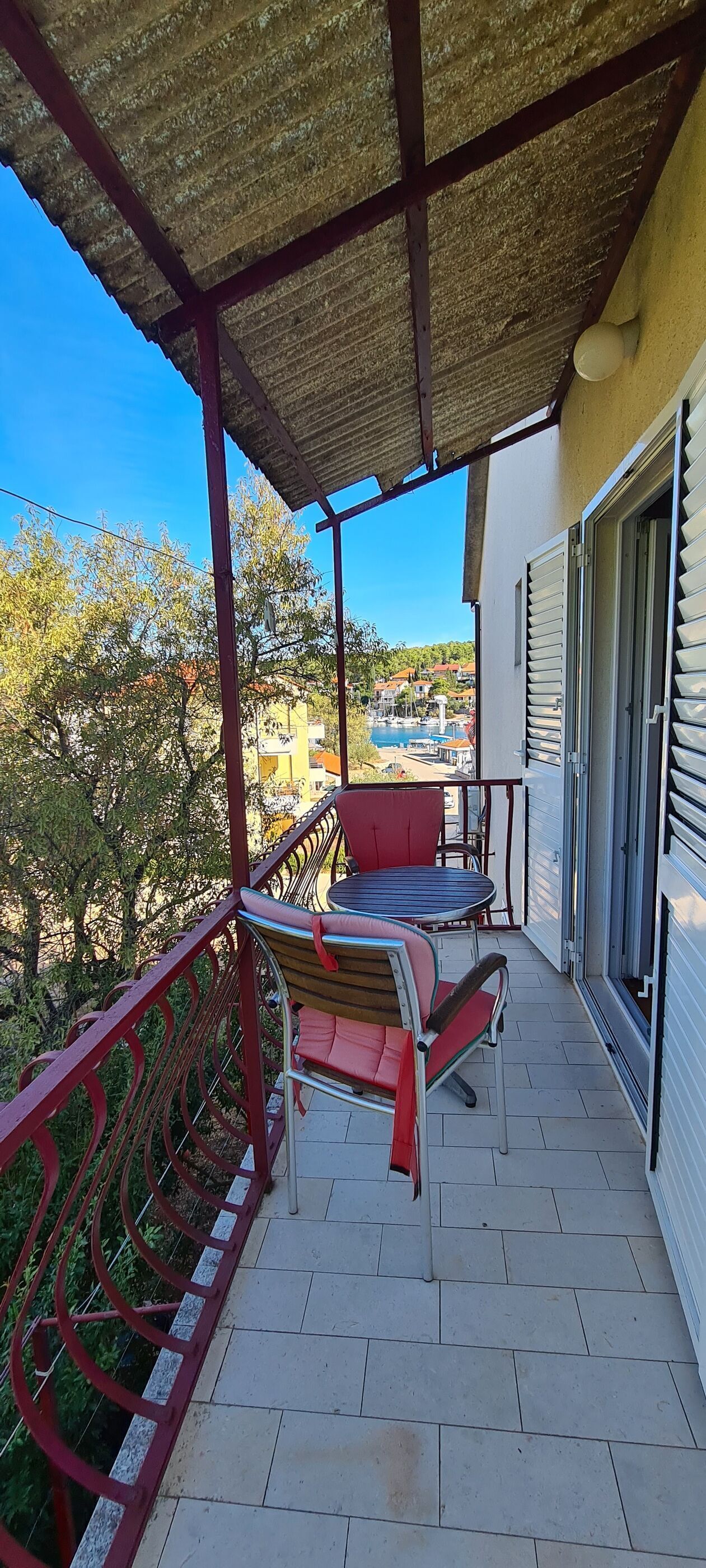 A covered balcony with a table and chairs, a view of a pool, and a building with a yellow facade.