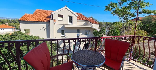 A balcony with red chairs and a small round table, overlooking a house with a red-tiled roof.