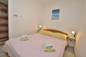 Un lit bien fait, avec une literie rose et deux serviettes enroulées, une table de chevet avec une lampe et un tableau représentant un paysage accroché au mur.