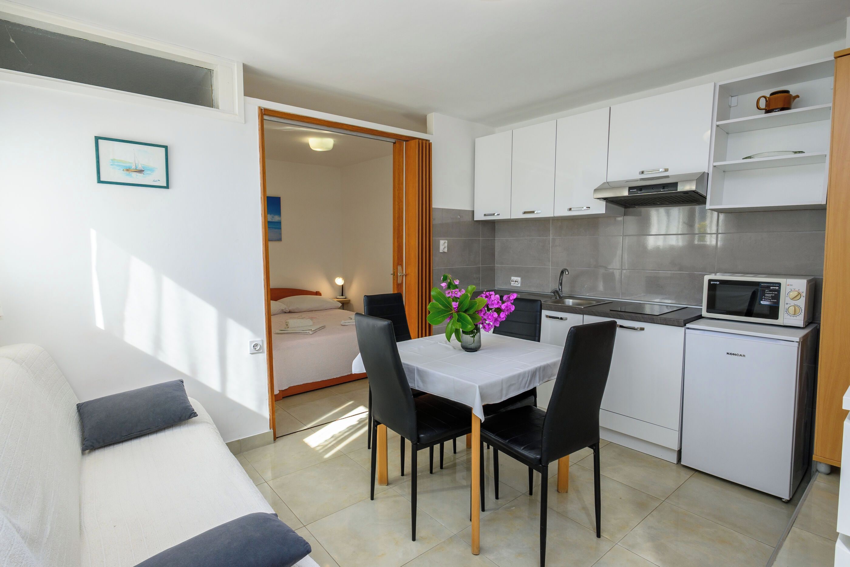 A compact kitchenette with a microwave, a sink, and a small refrigerator. There is a dining area with a table and chairs, and a bed is visible in the adjacent room.