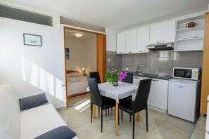 A compact kitchenette with a microwave, a sink, and a small refrigerator. There is a dining area with a table and chairs, and a bed is visible in the adjacent room. - Apartments Magnolia (Rabac)