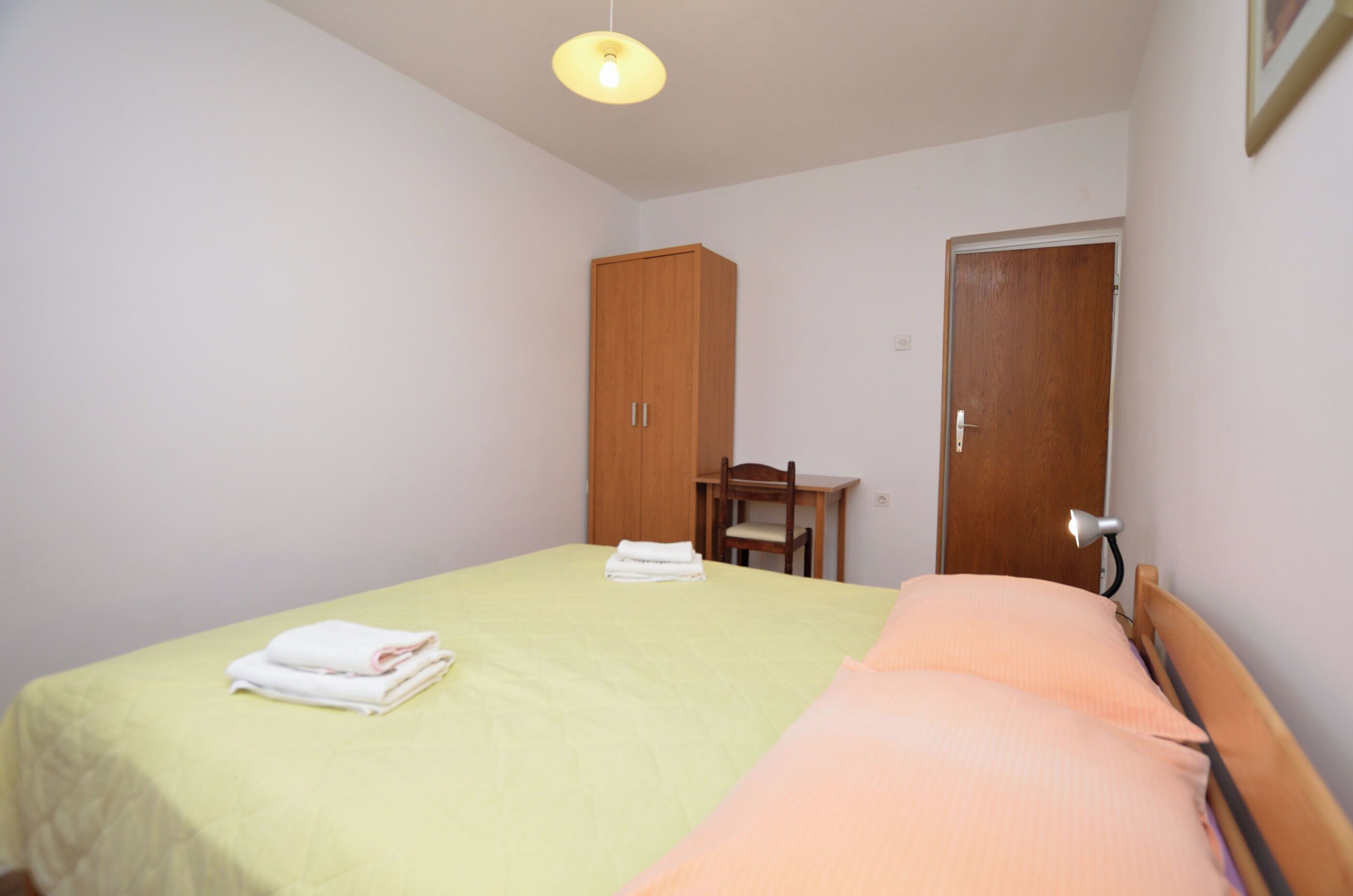 A small, clean room with a bed, a wooden wardrobe, a desk, and a door.