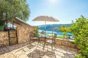 Une terrasse avec un sol en pierre, une table entourée de chaises, un grand parasol et une vue sur un plan d’eau et des collines.