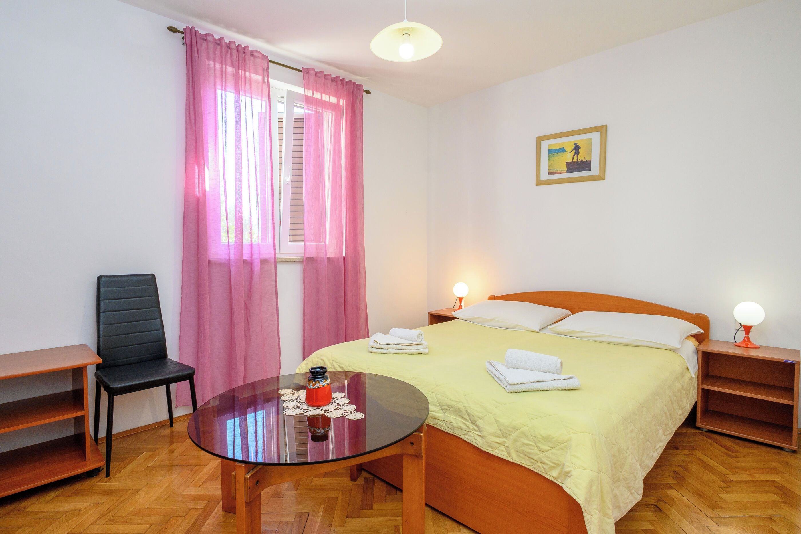 Habitación de hotel con cama, mesitas de noche, una silla, una mesa redonda con cubierta de vidrio y una ventana con cortinas.