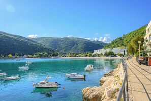 A coastal scene with boats docked along the shore, a beach, and a mountainous backdrop. - Apartments Magnolia (Rabac)