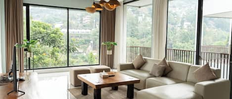 A modern living room with a sofa, a coffee table, and a TV. There are large windows with curtains and a view of greenery outside.