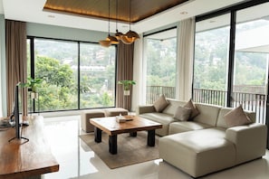 Un salon moderne avec un canapé, une table basse et un téléviseur. De grandes fenêtres, ornées de rideaux, offrent une vue sur la verdure environnante.