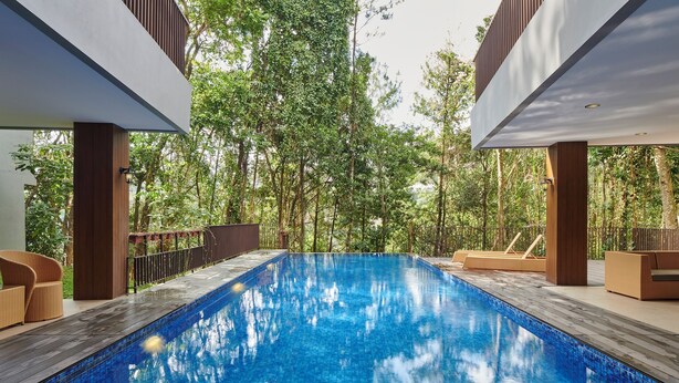 Una piscina con borde de azulejos, rodeada de mobiliario y sillones de madera, con árboles y una estructura de edificio moderno de fondo.