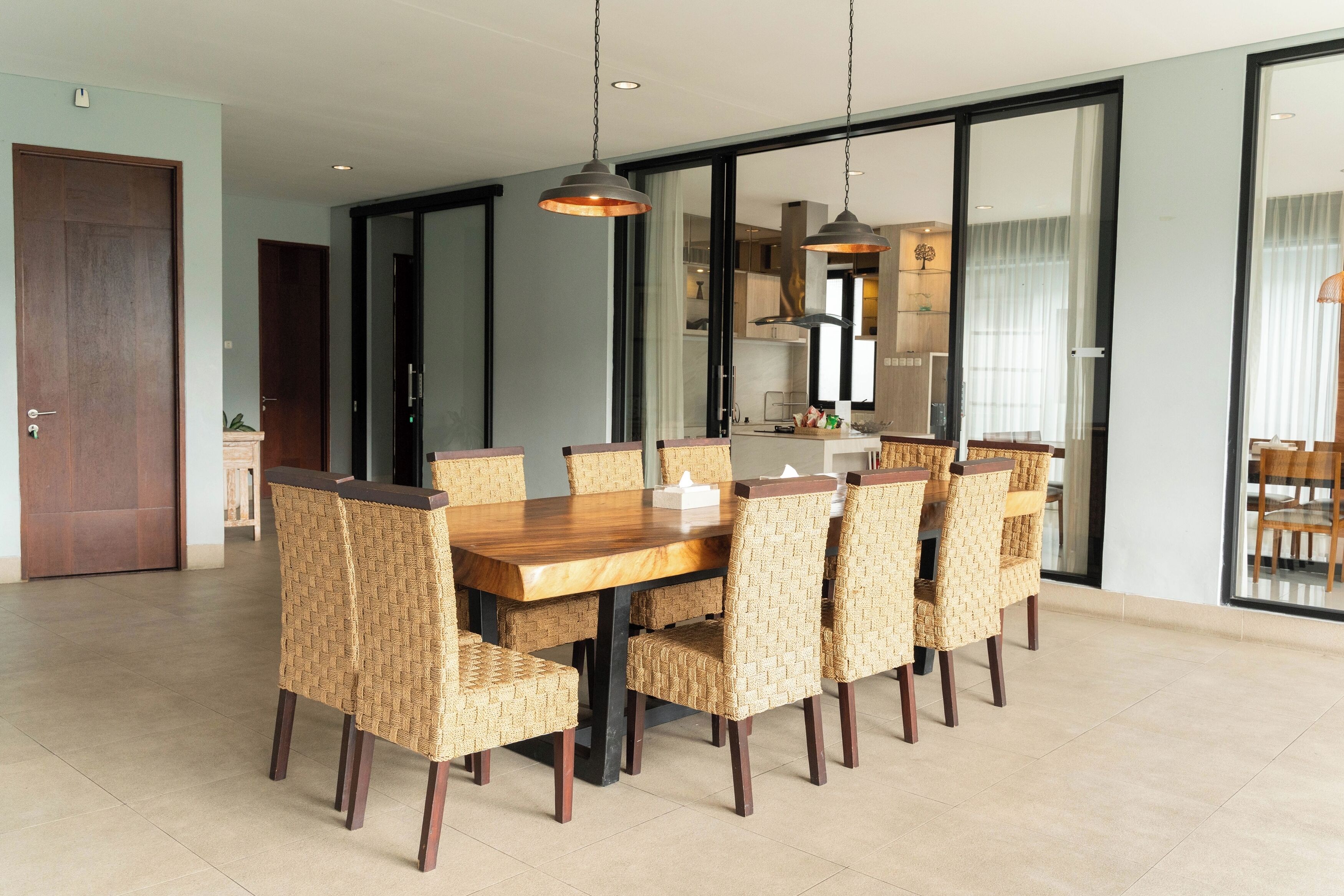 A dining area with a wooden table and woven chairs, a kitchen in the background, and a sliding glass door leading to another room.
