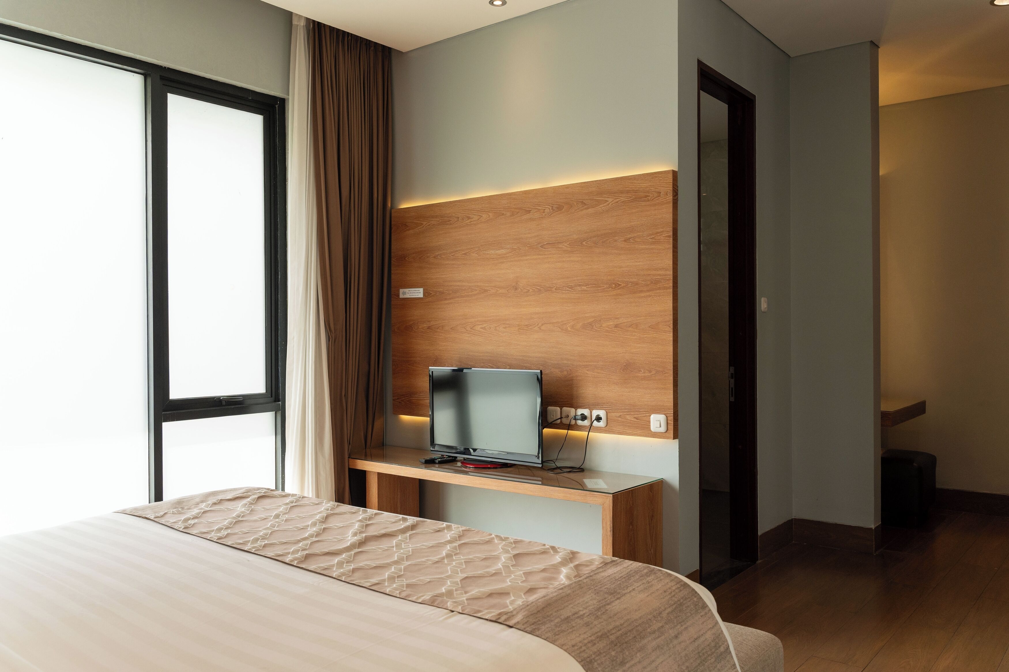 A hotel room with a bed, a TV on a wooden stand, a window with curtains, and a door.