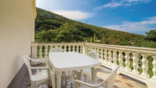 A balcony with a white table and chairs, a white balustrade, and a view of a green hillside. - Apartments Vukelic (Senj)
