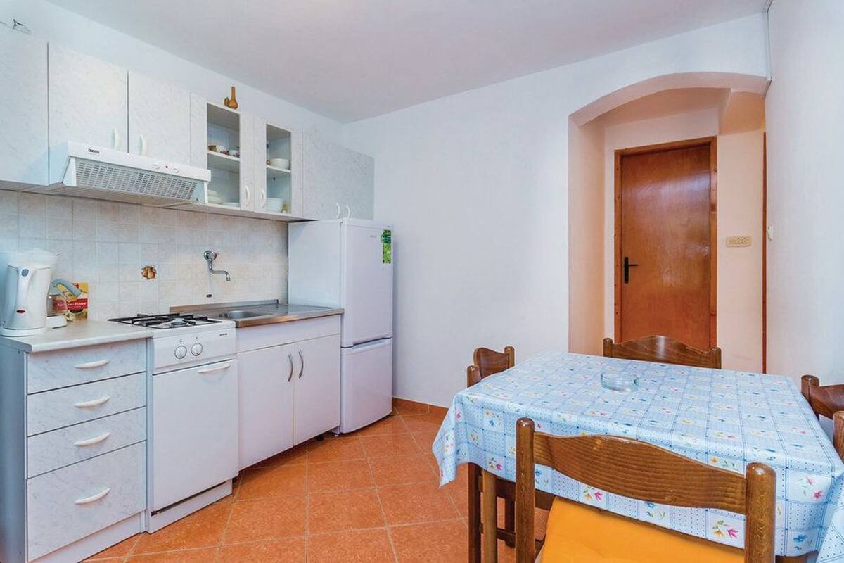 A compact kitchen with white cabinets, a stove, and a refrigerator. There is a dining table with a floral tablecloth and wooden chairs.