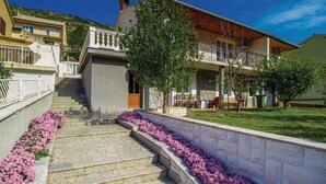 A two-story house with a stone staircase leading to a wooden door, surrounded by a garden with purple flowers. - Apartments Vukelic (Senj)