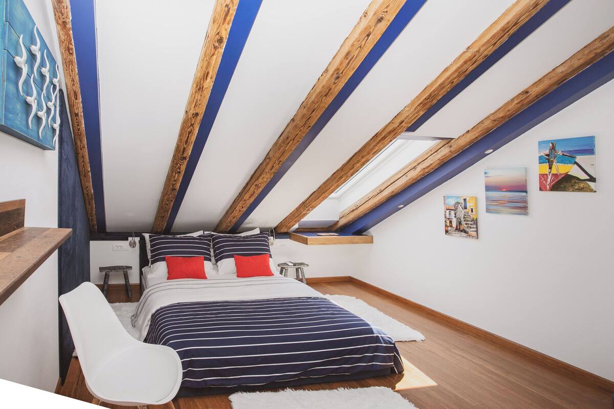 A bedroom with a sloped ceiling, wooden beams, a bed with striped bedding, a white chair, and artwork on the walls.