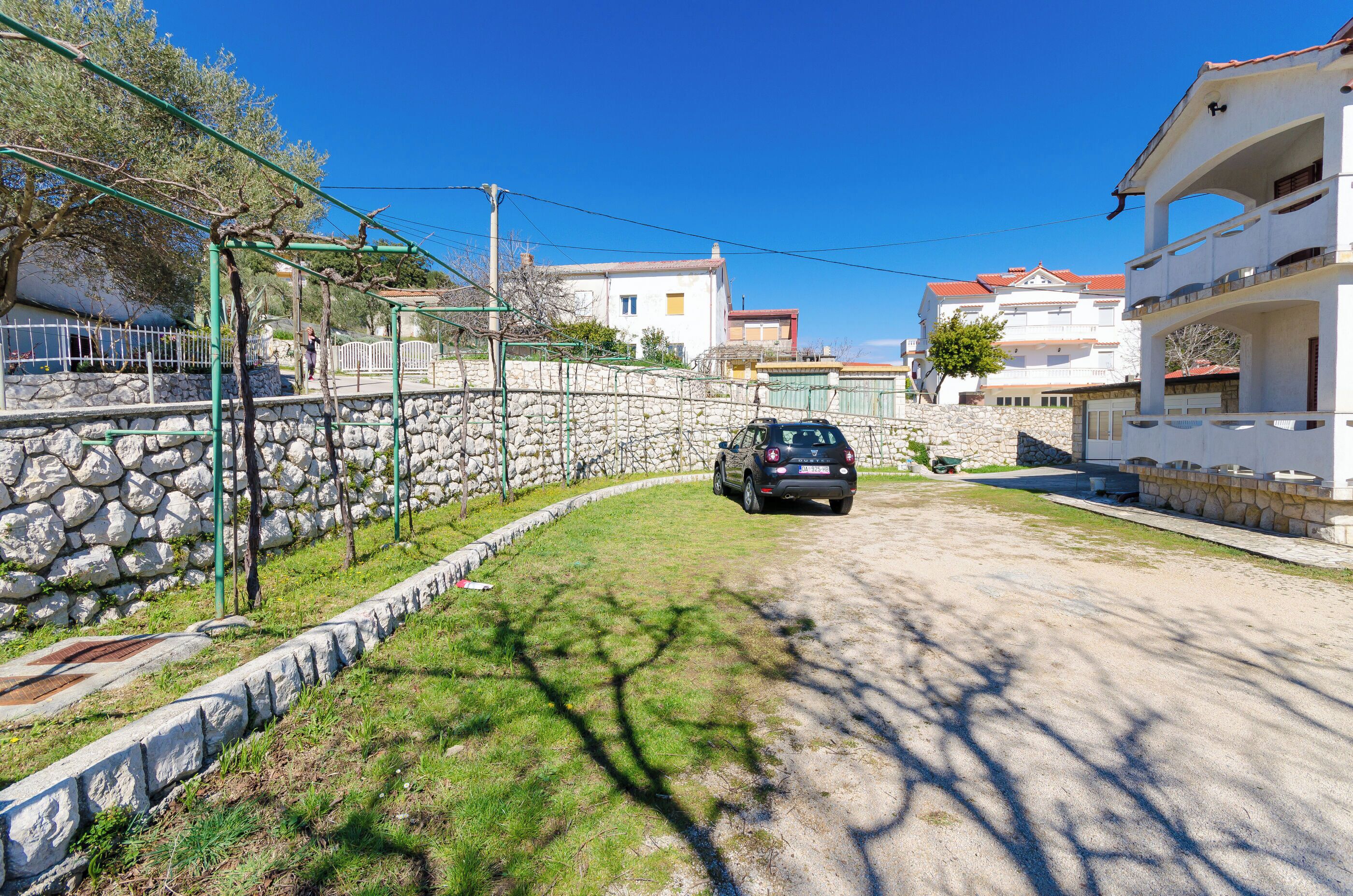 A residential street with a car parked on the side, stone walls, and houses with balconies.
