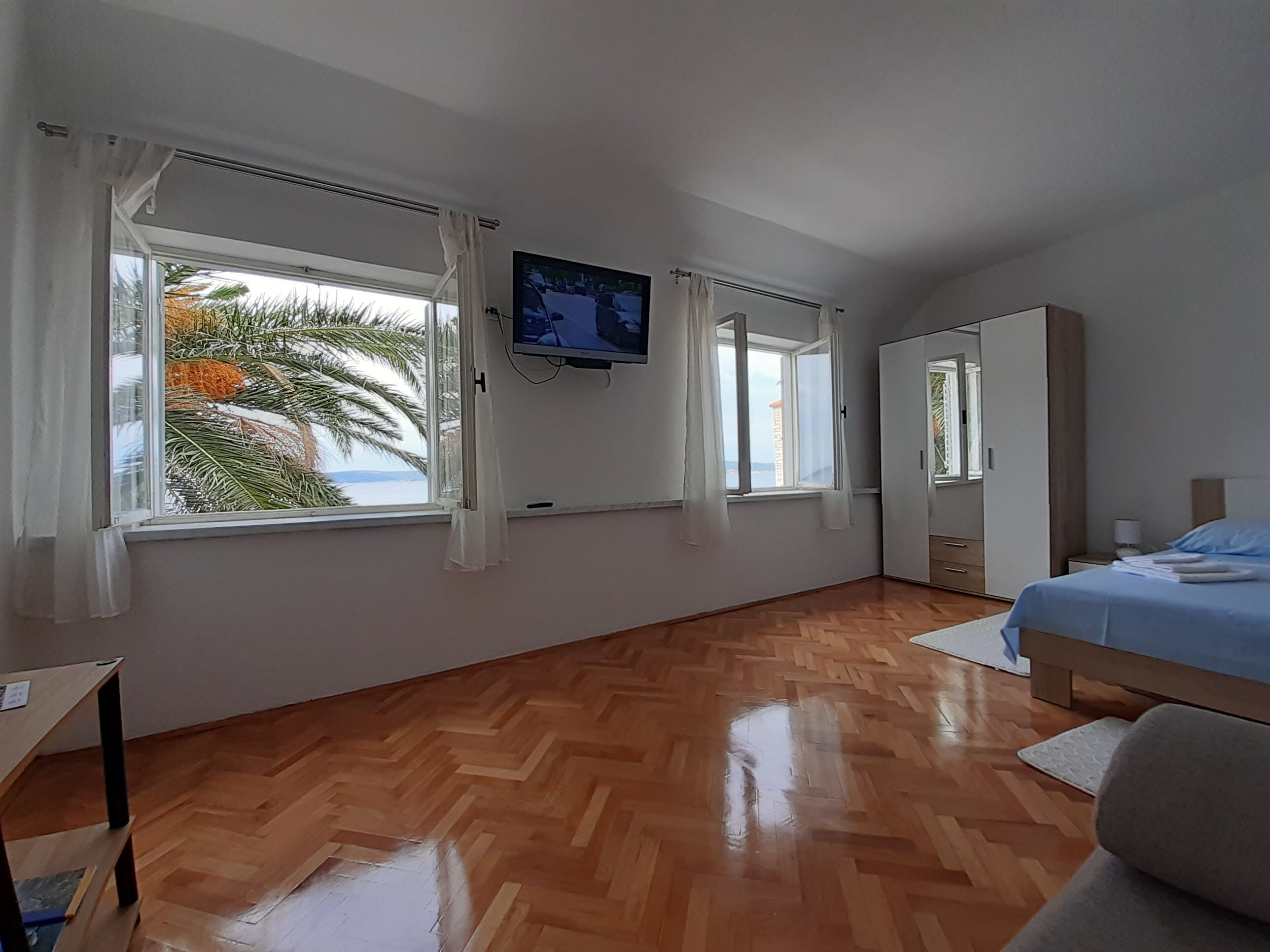 Un dormitorio con cama, televisor montado en la pared, armario y dos ventanas con cortinas.