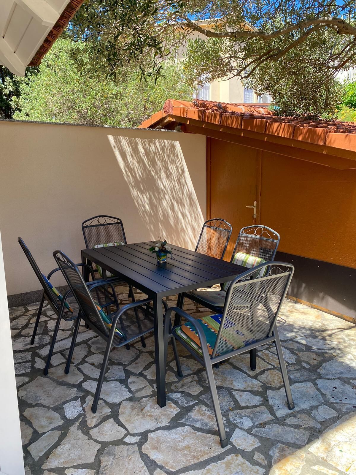 An outdoor patio with a table and chairs, stone flooring, and a view of trees and a building in the background.