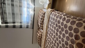 Ein Schlafzimmer mit einem Bett, das mit einer gemusterten Bettdecke bezogen ist, einem Fenster mit vertikalen Jalousien und einem Holzschrank.