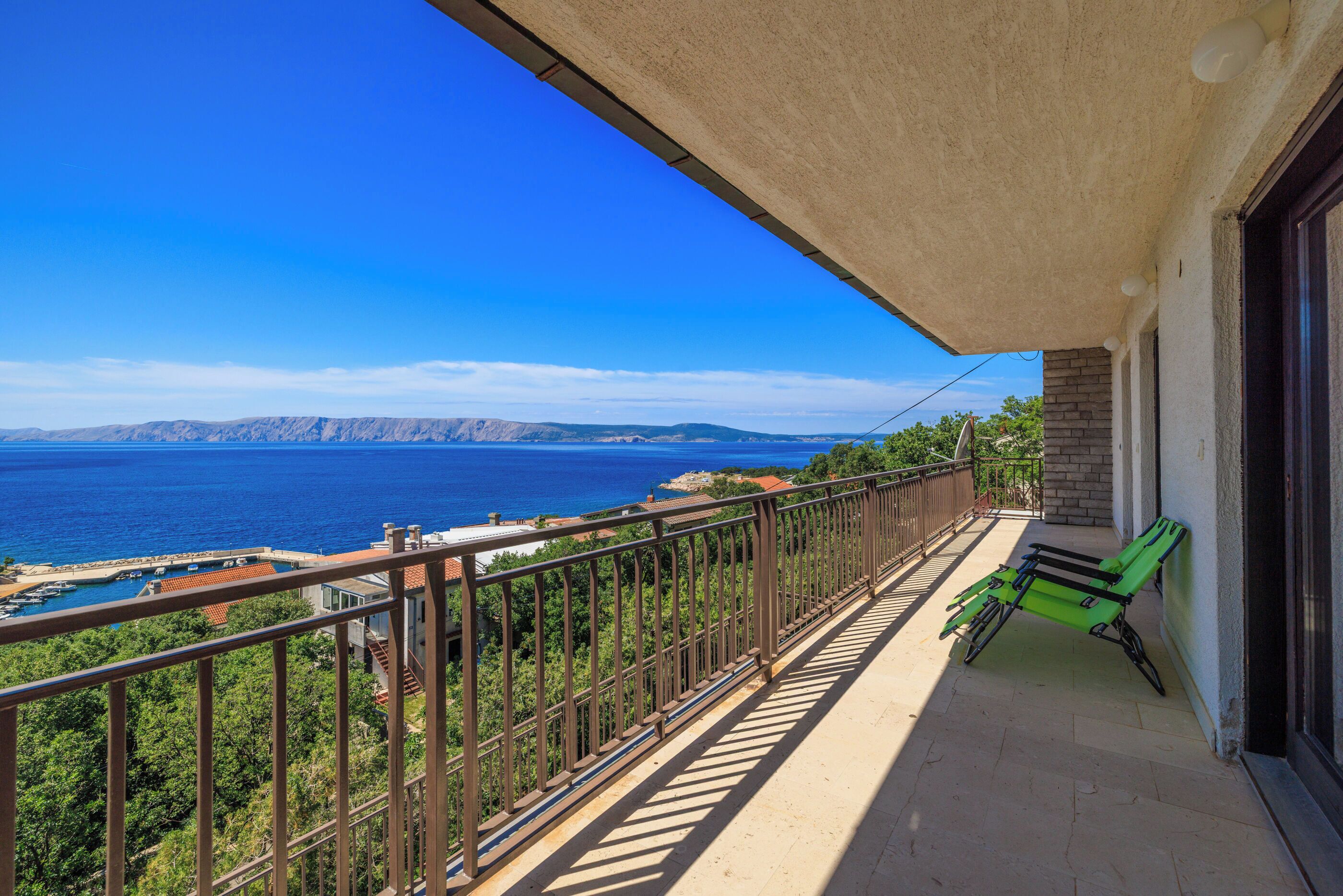 Een balkon met uitzicht op zee en bergen, een groene stoel en een balustrade.