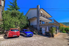 Una casa a due piani con un balcone, un vialetto e un'auto rossa e blu parcheggiata davanti.