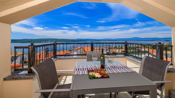 A balcony with a table set for two, a bottle of wine, and a plate of food. The balcony overlooks a scenic view of a body of water and distant mountains. - Apartments Jozic (Crikvenica)