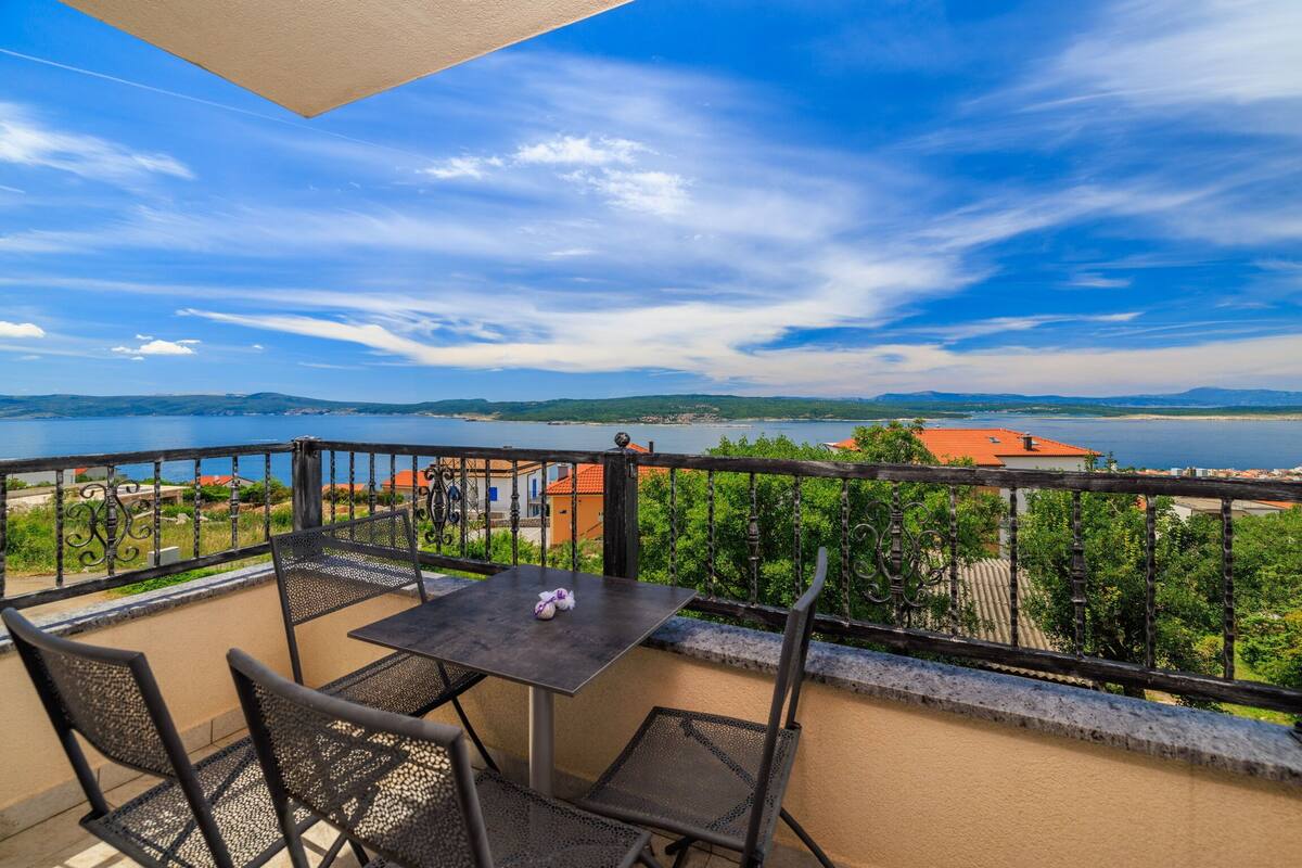 A balcony with a table and chairs, overlooking a body of water and distant hills.