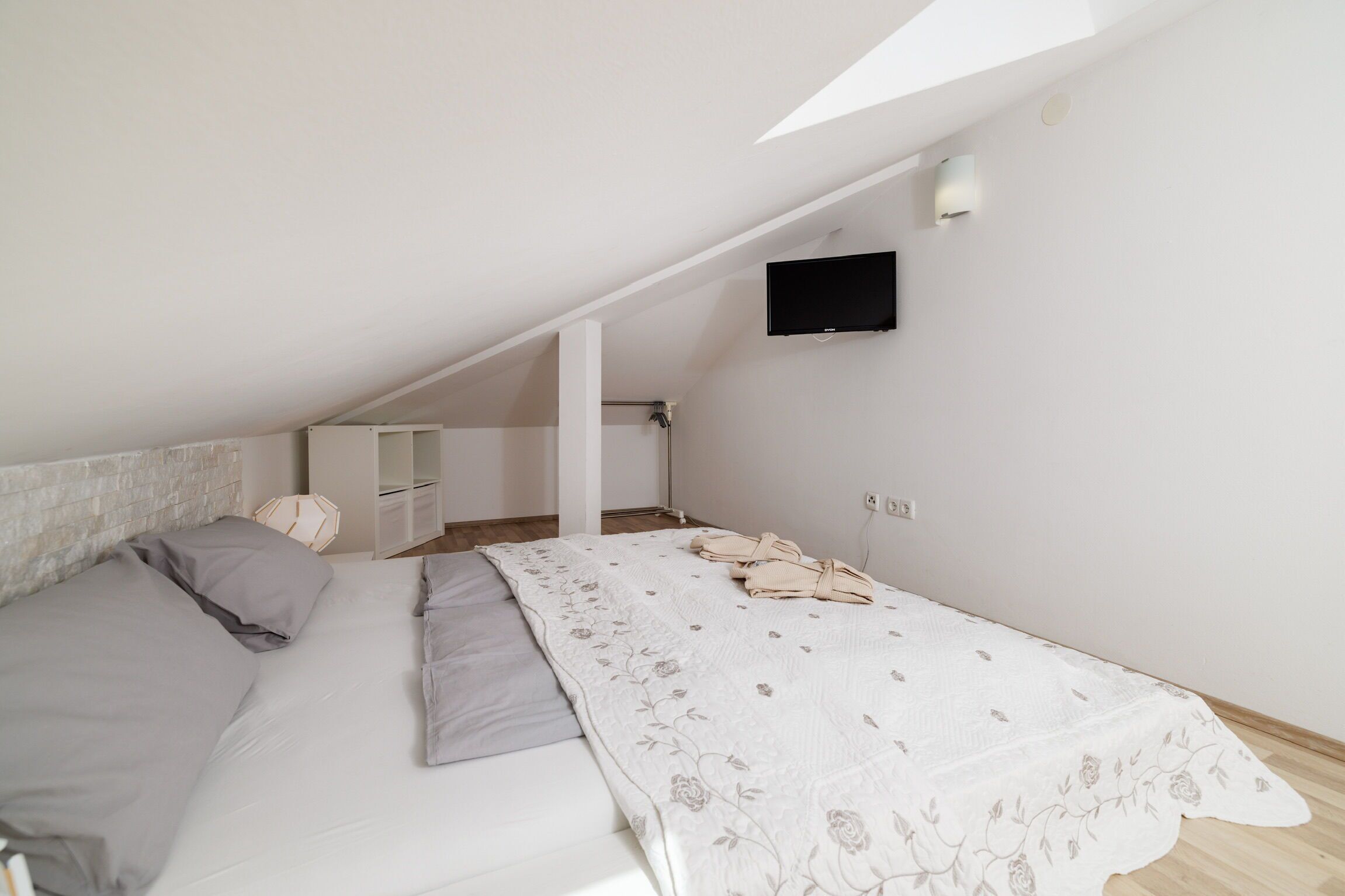 A bedroom with a sloped ceiling, a bed with grey bedding, a white wall-mounted TV, and a small bedside table.