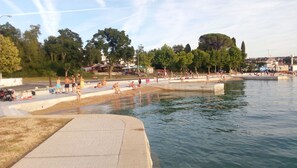 Um parque à beira-mar com pessoas relaxando à beira da água, um calçadão de concreto e árvores ao fundo.