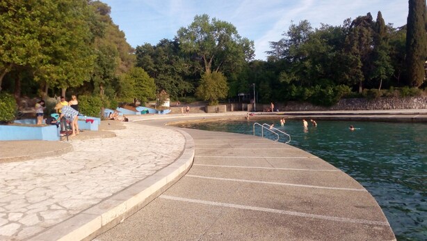 Ein Schwimmbad mit einem steinbelagten Weg zum Wasser, umgeben von Bäumen und einer Steinmauer.