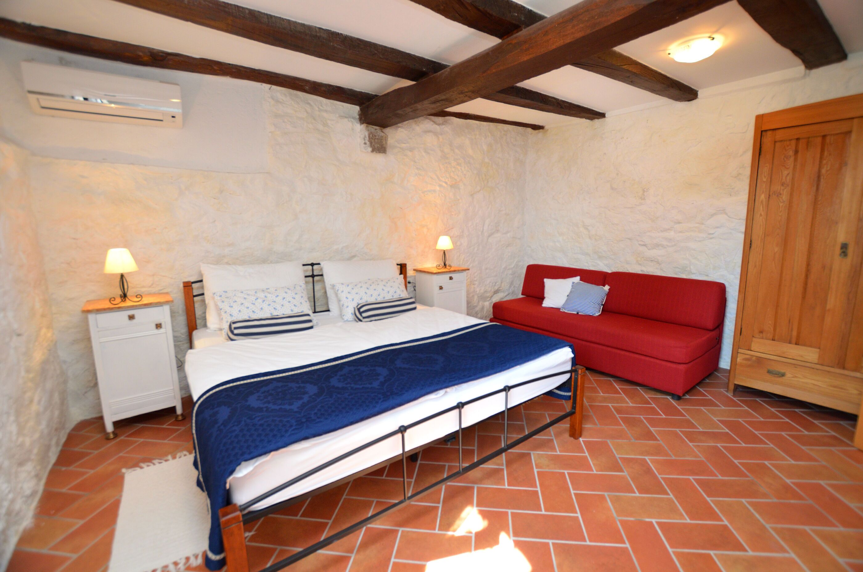 A bedroom with a bed, bedside tables, a red sofa, a wooden wardrobe, and a tiled floor.