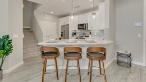 Een moderne keuken met een centraal eiland, witte kasten, roestvrijstalen apparatuur en drie houten barstoelen.