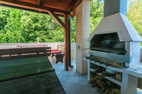 An outdoor patio with a wooden table and benches, a barbecue grill, and a view of a forested area. - Apartments Ljiljana (Supetarska Draga)
