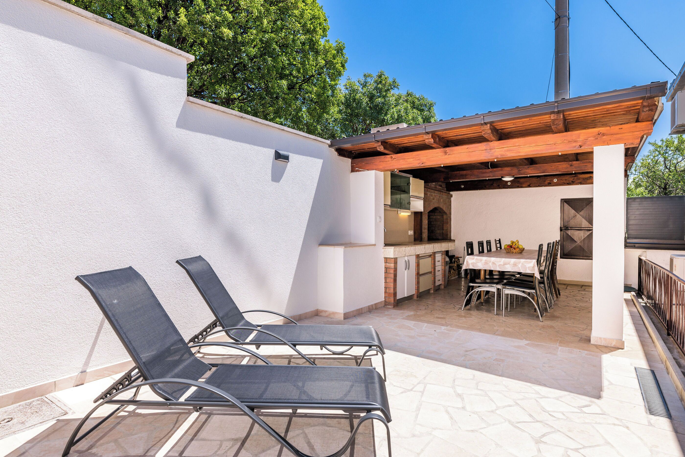 An outdoor patio with a pergola, a dining table with chairs, and a kitchen area.