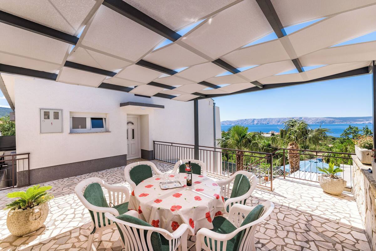 A covered outdoor dining area with a table and chairs, a view of the sea, and a stone-tiled floor.