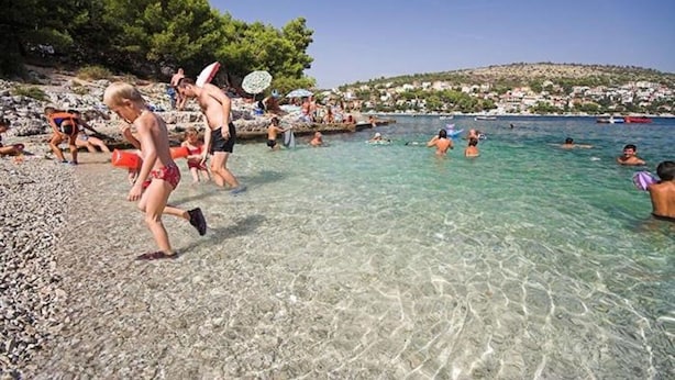 A beach scene with clear water, people swimming and walking along the shore, and a hillside in the background. - Apartments Kursan (Okrug Gornji)
