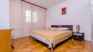 A bedroom with a wooden floor, a bed with a beige blanket, a nightstand with a lamp, and a window with sheer curtains. - Apartments Kursan (Okrug Gornji)