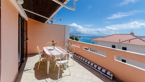 A balcony with a table and chairs, a view of the sea, and a building with red-tiled roofs. - Apartments Kursan (Okrug Gornji)