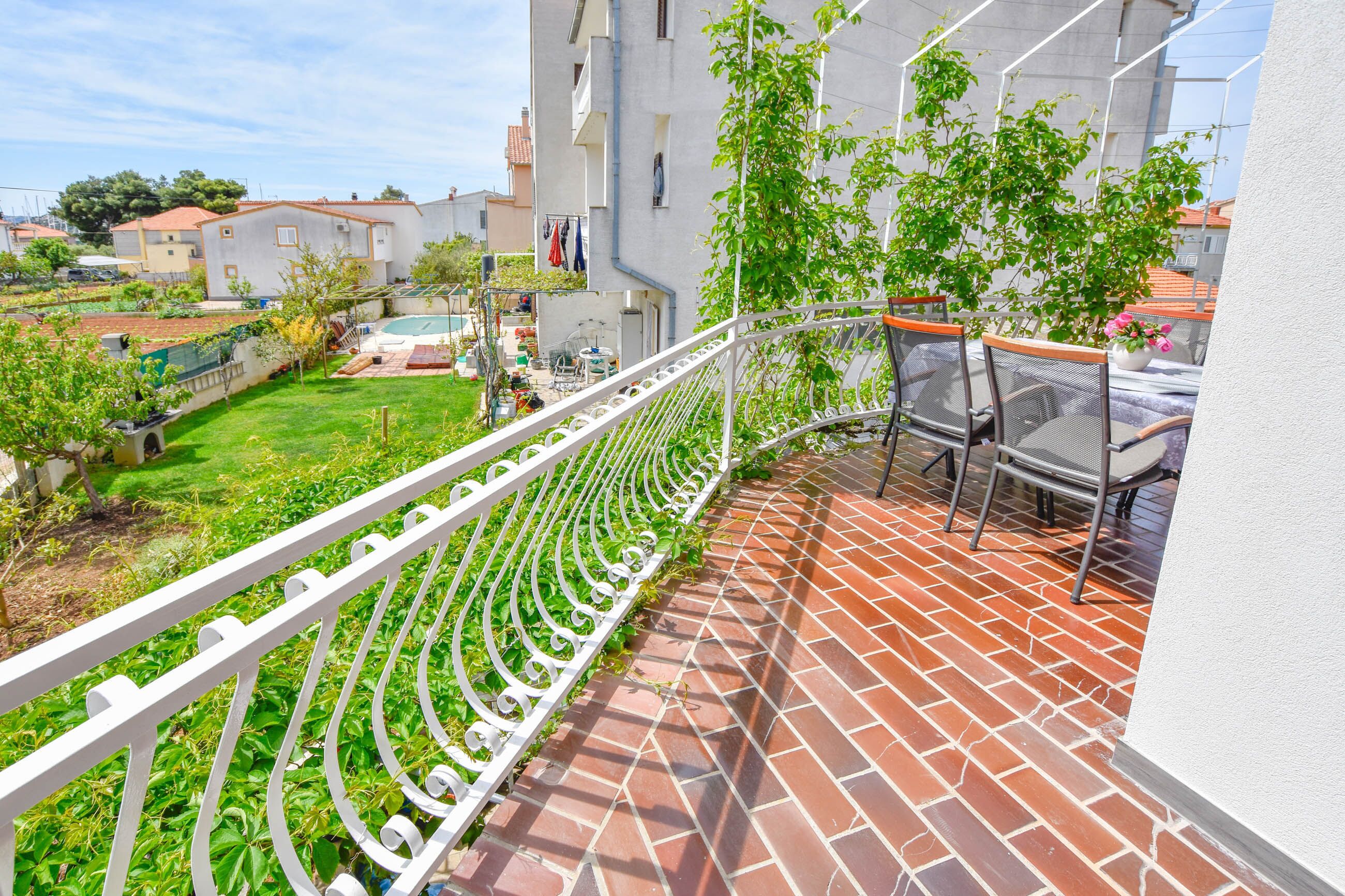 A balcony with a white metal railing, a brick floor, and a view of a garden and neighboring buildings.