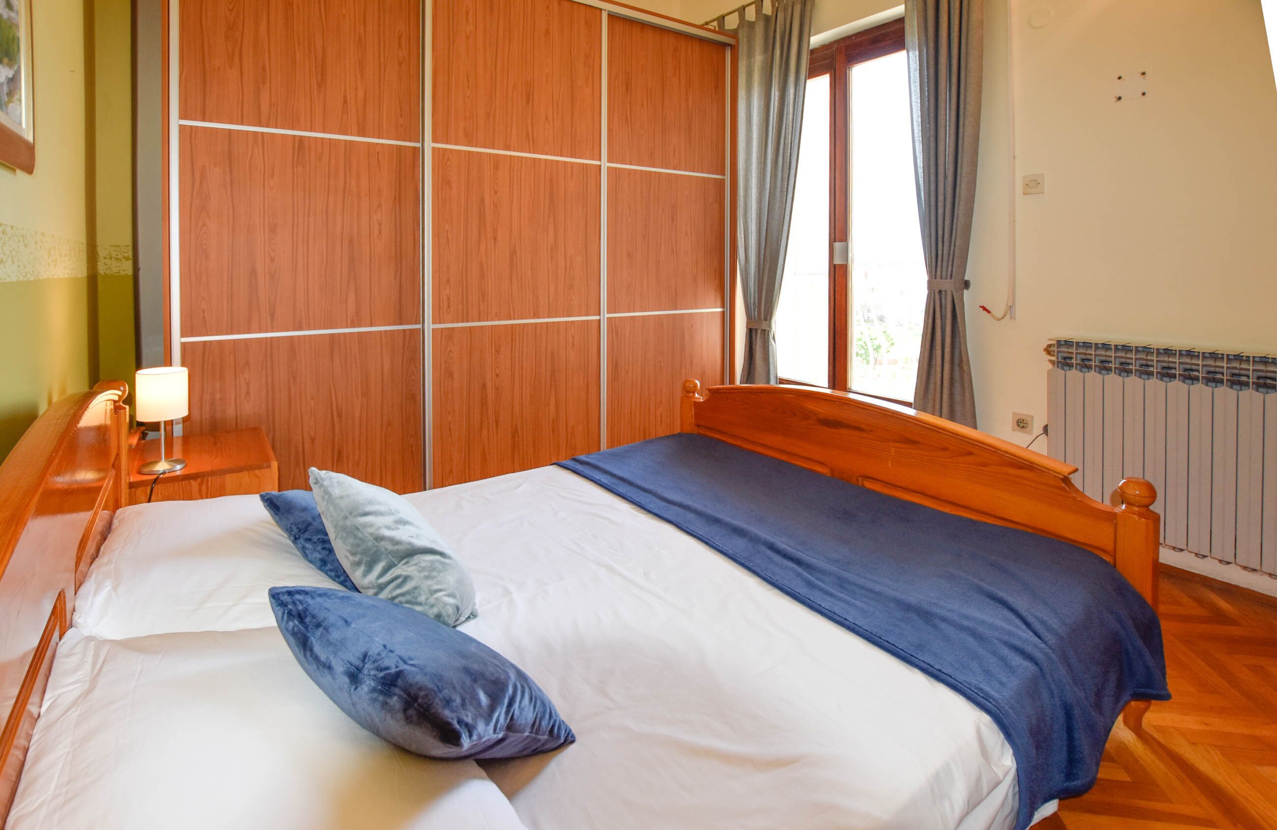 A bedroom with a wooden bed, a nightstand with a lamp, and a view of the outdoors through a window.