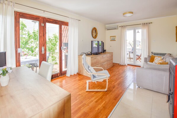 A spacious living room with a wooden floor, a dining table, a sofa, a TV, and a window with curtains.