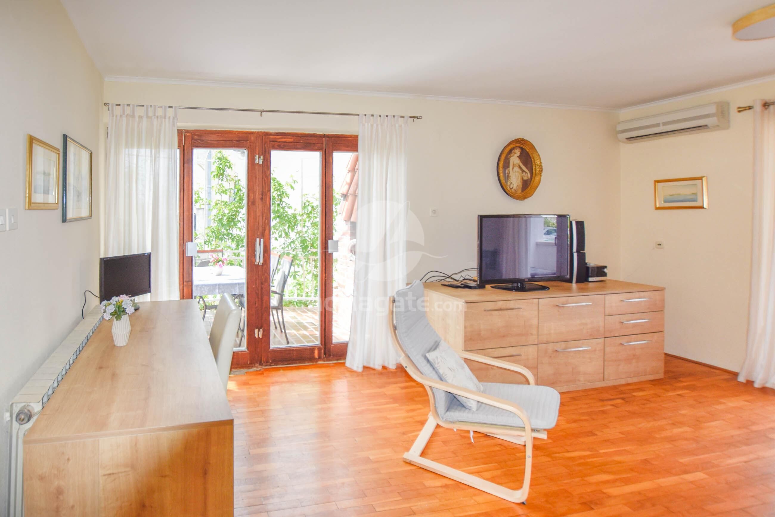 Une pièce dotée d’un plancher en bois, d’un bureau avec un ordinateur, d’une télévision, d’une chaise et d’une porte-fenêtre coulissante donnant sur un espace extérieur agrémenté de plantes.